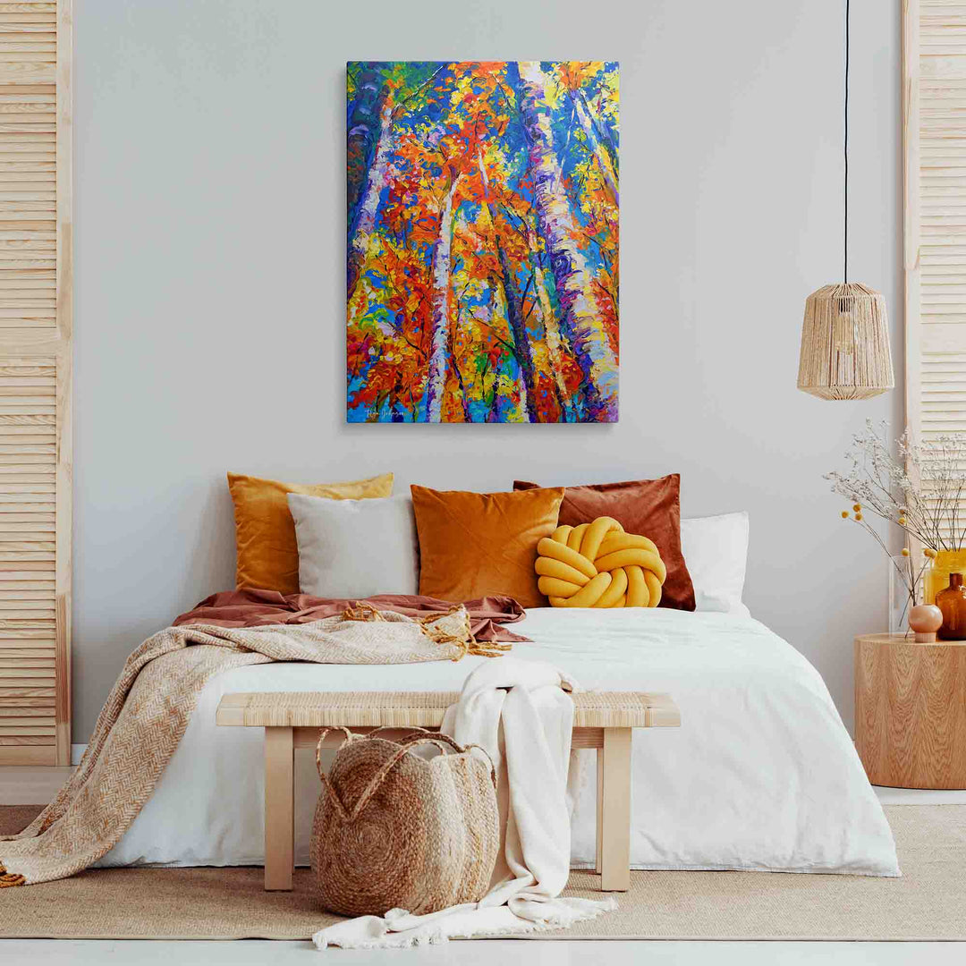 A colorful abstract canvas print of autumn trees hangs on a light gray wall above a styled bed with orange, yellow, and white pillows, a wooden bench, and neutral-toned decor in a bright bedroom setting.