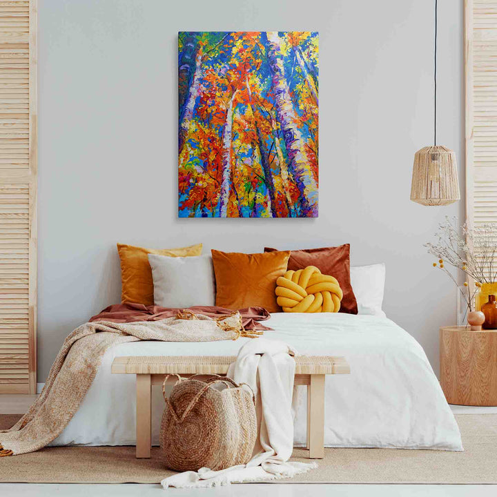 A colorful abstract canvas print of autumn trees hangs on a light gray wall above a styled bed with orange, yellow, and white pillows, a wooden bench, and neutral-toned decor in a bright bedroom setting.