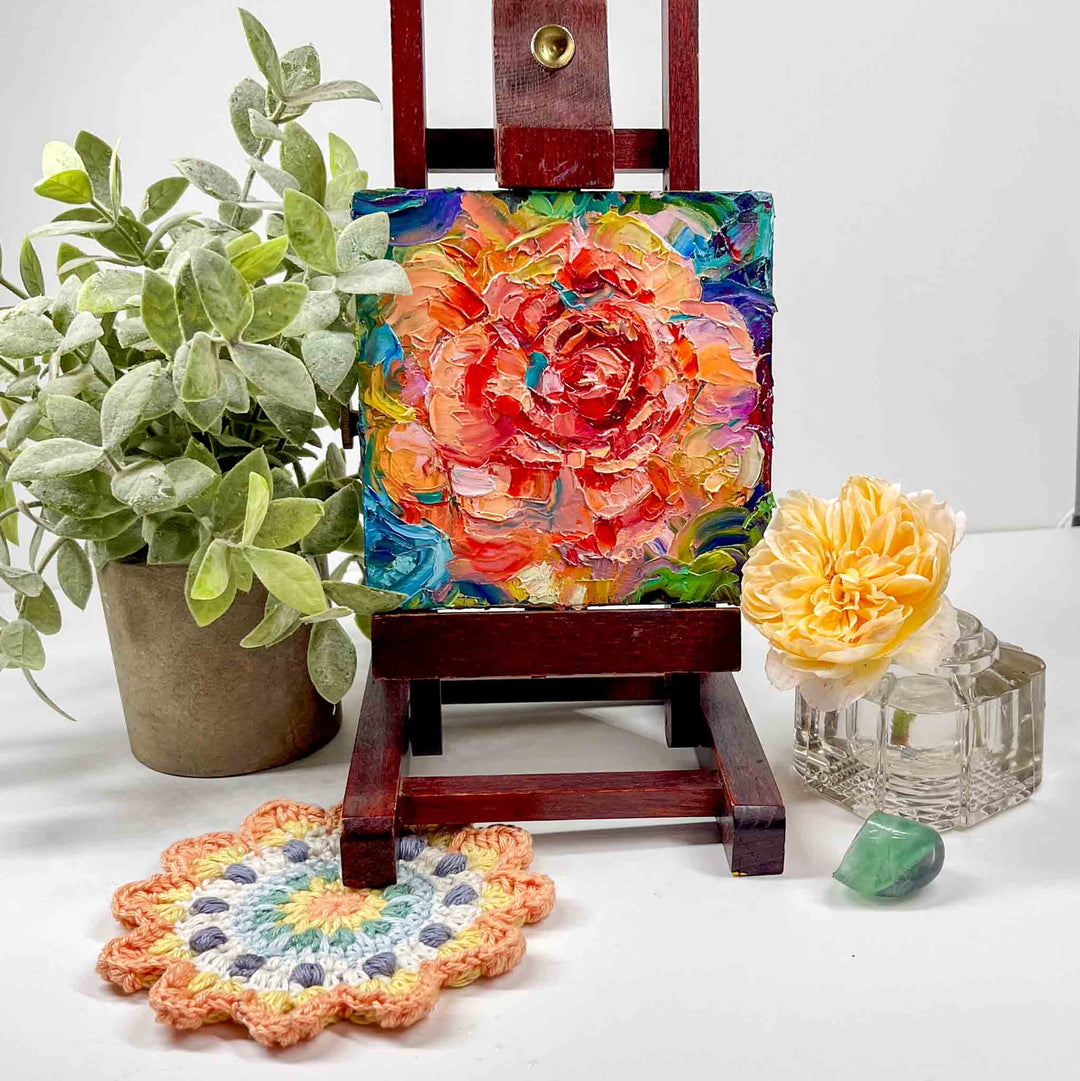 Palette knife original oil painting depicting an abstract orange rose with blue green modern background. Impasto painting by Oregon artist Talya Johnson. Mini painting is displayed on a small easel placed on a shelf with natural decor.