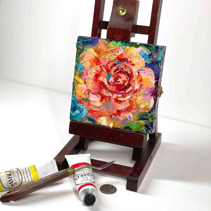 Palette knife original oil painting depicting an abstract orange rose with blue green modern background. Impasto painting by Oregon artist Talya Johnson. Mini painting is displayed on a small easel placed on a shelf with oil painting supplies.