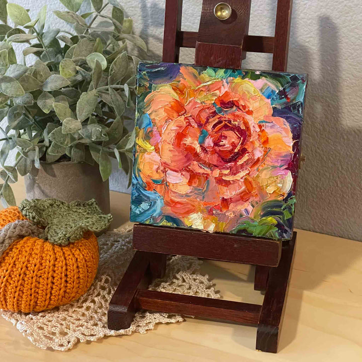 Impasto painting painted with palette knife depicting an abstracted orange rose with blue green background. Original oil painting is displayed on a small easel placed on a shelf with nature inspired fall decor.